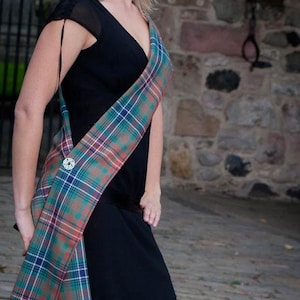 May include: A woman wearing a black dress with a plaid sash. The sash is tied around her waist and drapes down her side. She is standing in front of a stone wall.