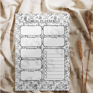 May include: A white meal planner with a black and white food-themed border. The planner includes sections for each day of the week and a grocery list. The words "Meal Planner" are at the top.