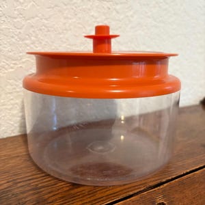 May include: A clear glass jar with an orange plastic lid and a small orange knob on top. The jar is empty.