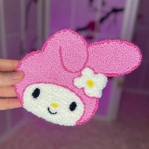 May include: Pink and white plush character with a bow and a flower. The character has a smiling face with black eyes and a yellow nose.