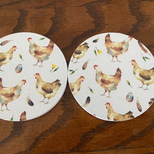 May include: Two round white coasters featuring a watercolour design of brown chickens, yellow flowers, green leaves, and blue eggs.