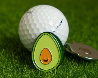 May include: A green avocado golf ball marker with a smiling face. The marker is attached to a silver golf ball retriever.