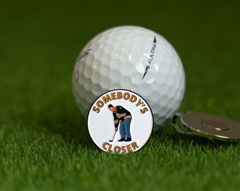 May include: A white golf ball with a black and white logo on it. A white golf ball marker with a cartoon of a hockey player and the text "Somebody's Closer" is in front of the golf ball.