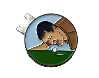 May include: A round, metal golf ball marker with a cartoon image of a man in a brown shirt, looking down at a golf ball on a green putting surface. The man is making a face of frustration.