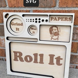 Könnte beinhalten: Ein Holz-Rolltablett mit Fächern, die mit "Grind It", "Wax It" und "Papers" beschriftet sind. Das Tablett hat auch ein großes Fach mit der Aufschrift "Roll It" und ein kleines Fach mit einem Cartoon-Bild einer Person, die raucht.