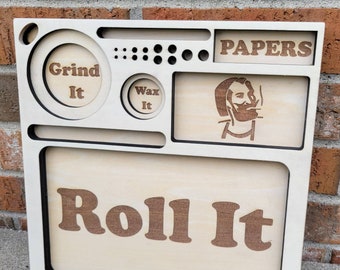 Könnte beinhalten: Ein Holz-Rolltablett mit Fächern, die mit "Grind It", "Wax It" und "Papers" beschriftet sind. Das Tablett hat auch ein großes Fach mit der Aufschrift "Roll It" und ein kleines Fach mit einem Cartoon-Bild einer Person, die raucht.