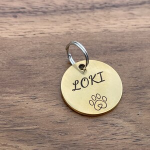 Op de afbeelding: Een gouden metalen hondenpenning met een zilveren ring. De penning is gegraveerd met de naam "LOKI" en een pootprint met een hart erin.