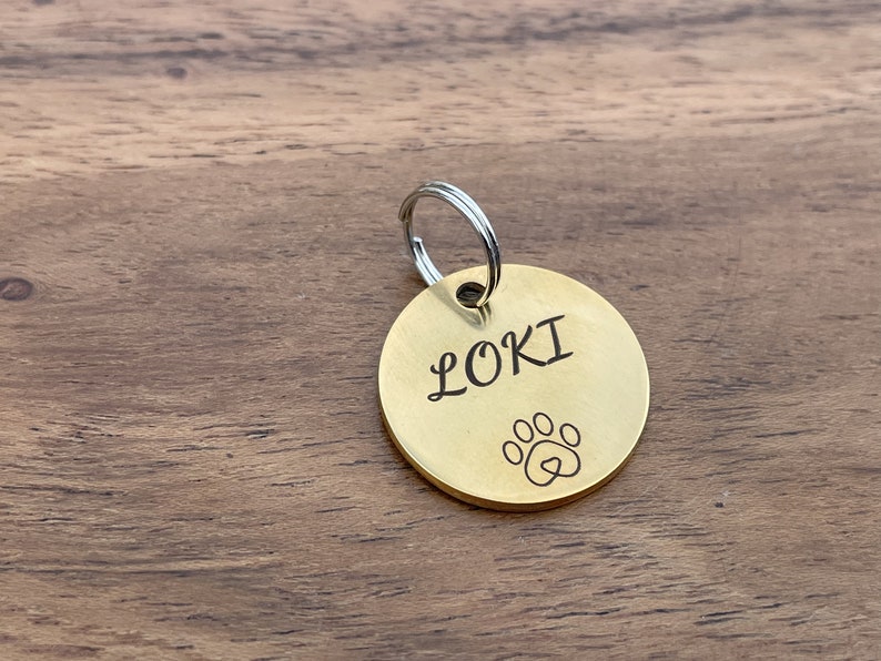 Op de afbeelding: Een gouden metalen hondenpenning met een zilveren ring. De penning is gegraveerd met de naam "LOKI" en een pootprint met een hart erin.