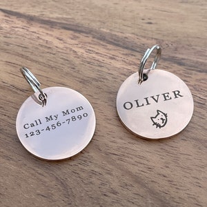 Op de afbeelding: Twee ronde, zilverkleurige huisdier-ID-tags met gegraveerde tekst. Een tag luidt "Call My Mom 123-456-7890" en de andere tag luidt "OLIVER" met een klein kattenicoon.