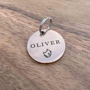 Op de afbeelding: Een ronde, zilveren huisdiertag met de naam "OLIVER" erop gegraveerd. Een klein katten silhouet is gegraveerd onder de naam.