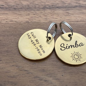 Op de afbeelding: Twee gouden hondenpenningen met een zilveren sleutelring. Een penning zegt "Call My Mom 123-456-7890" en de andere penning zegt "Simba" met een zonnetje.