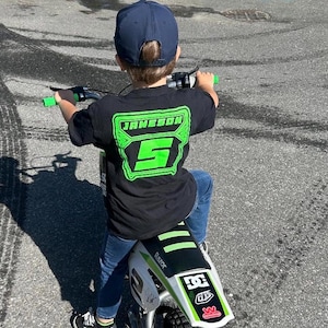 May include: A child wearing a black t-shirt with the name "JAMESON" and the number "5" in neon green, riding a white and green electric motorcycle. The child is wearing a navy blue baseball cap and blue jeans.