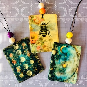 May include: Three rectangular wooden ornaments hanging from black cords with colorful wooden beads. One features a bee and flowers on a yellow background, one shows moon phases on a dark teal background, and one displays a sun and moon on a blue background.
