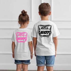 May include: Two white t-shirts featuring a graphic on the back. The smaller shirt has a pink outline of a shield shape with the text BELL 607. The larger shirt has a black outline of the same shield shape with the text BELL 607.