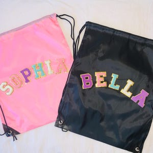 May include: Two drawstring bags, one bright pink and one black, each featuring large, glittery, multi-colored varsity-style letters. The pink bag displays the name SOPHIA, while the black bag displays the name BELLA. Both bags have black cords.