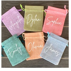 May include: Six small burlap drawstring bags in various colors with names written in white on the front. The names are: Angela, Dylan, Sophia, Emily, Olivia, and James.