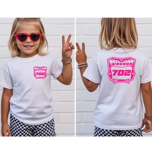 May include: A white short-sleeved t-shirt featuring a pink graphic on the front left chest and a larger matching pink graphic on the back. The graphic displays the text WAGNOR and the number 702 inside a stylized shield-like border design.