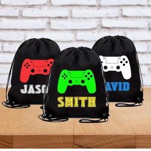 May include: Three black drawstring bags featuring a video game controller graphic. The left bag has a red controller and the name JASON in white. The center bag has a green controller and the name SMITH in yellow. The right bag has a white controller and the name DAVID in blue.