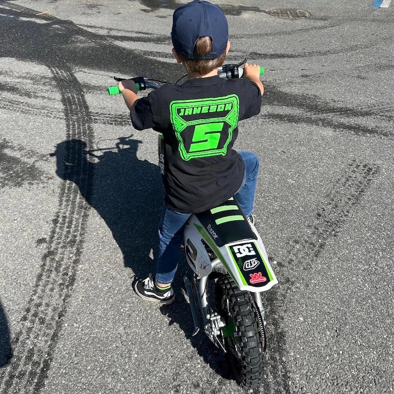 May include: A child wearing a black t-shirt with the name "JAMESON" and the number "5" in neon green, riding a white and green electric motorcycle. The child is wearing a navy blue baseball cap and blue jeans.