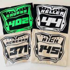 May include: Four t-shirts featuring a shield-shaped graphic with stars and custom text. One black shirt has neon green text reading GRUBBY 402, one black shirt has white text reading KELLER 44, one tan shirt has black text reading ACREMAN 371, and one tan shirt has black text reading NICK 145.