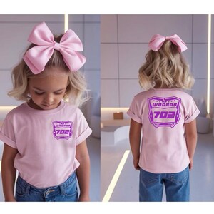 May include: A light pink t-shirt featuring a purple graphic on the front chest and a larger matching purple graphic on the back. The graphic displays the text WAGNOR and the number 702 inside a shield shape. A large light pink hair bow is worn.