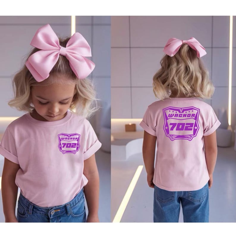 May include: A light pink t-shirt featuring a purple graphic on the front chest and a larger matching purple graphic on the back. The graphic displays the text WAGNOR and the number 702 inside a shield shape. A large light pink hair bow is worn.