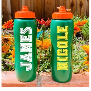 May include: Two green plastic water bottles with orange screw-top lids. The left bottle features the name JAMES in white block letters, while the right bottle features the name NICOLE in yellow block letters. Both bottles have a vertical orientation.