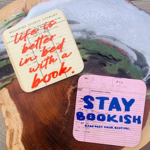 May include: Two square coasters featuring vintage library checkout card designs. The cream coaster has red cursive text reading "life is better in bed with a book." The pink coaster has blue block text reading "STAY BOOKISH" and "READ PAST YOUR BEDTIME."