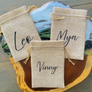May include: Three rectangular tan burlap drawstring pouches featuring black script lettering. The left pouch displays the name Leo, the top right pouch displays the name Mya, and the bottom center pouch displays the name Vinny in a cursive font.