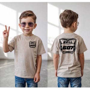 May include: A beige short-sleeve t-shirt featuring a black graphic on the front left chest and a larger matching black graphic on the back. The graphic displays the text BELL 607 inside a shield-like emblem with small stars at the top.