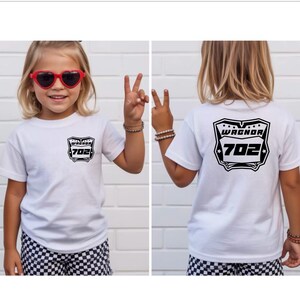 May include: A white short-sleeve t-shirt featuring a black graphic on the front left chest and a larger version of the same graphic on the back. The graphic displays a shield-like emblem with the text WAGNOR and the number 702 inside.