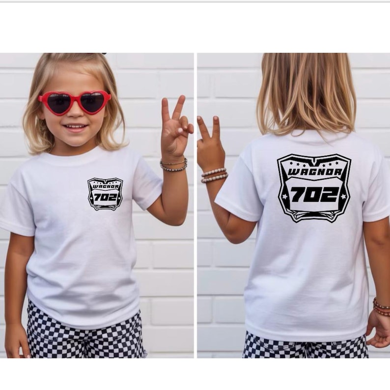 May include: A white short-sleeve t-shirt featuring a black graphic on the front left chest and a larger version of the same graphic on the back. The graphic displays a shield-like emblem with the text WAGNOR and the number 702 inside.