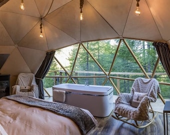 May include: A white bathtub sits in a geodesic dome with a large window overlooking a forest. The dome has a wooden interior with a bed, two chairs, and a small table with a lamp.