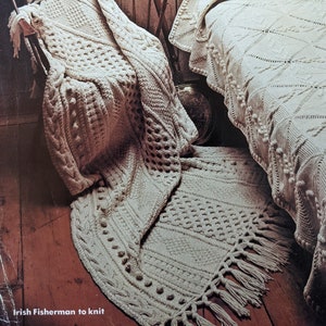 May include: A white crocheted throw blanket with a cable knit pattern and fringe. The blanket is draped over a wooden chair and a bed with a matching blanket.