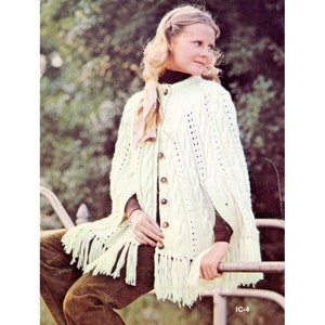 Op de afbeelding: Een witte gebreide poncho met een kabelbreipatroon en franjes. De poncho heeft een knoopsluiting en een sjaal die om de nek is geknoopt.