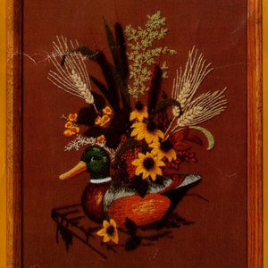 Puede incluir: Imagen bordada de un pato silvestre macho con plumaje marrón, naranja y verde, sentado en un jarrón de flores amarillas y tallos de trigo. El fondo es marrón oscuro.