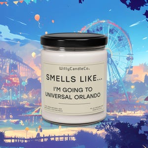 May include: A white candle jar with a black lid and a label that reads "Witty Candle Co. Smells Like... I'm Going To Universal Orlando". The candle is sitting on a table in front of a colorful cartoon illustration of a theme park.