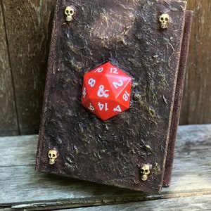 May include: A red 20-sided die with white numbers sits on top of a brown leather-bound book with gold skull accents.