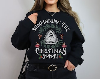 May include: Black sweatshirt with the text "SUMMONING THE CHRISTMAS SPIRIT" in white script. The design includes a planchette, holly, candy canes, and mugs of hot cocoa. The sweatshirt is a festive holiday themed item.