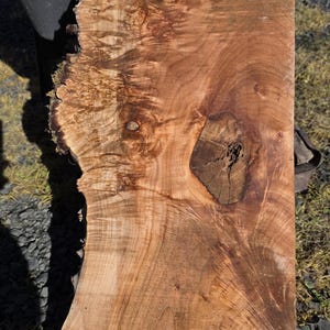 May include: A rectangular, unfinished slab of wood with a natural edge. The wood grain features swirling patterns and a mix of light and dark brown tones. A knot and a crack are visible in the wood.