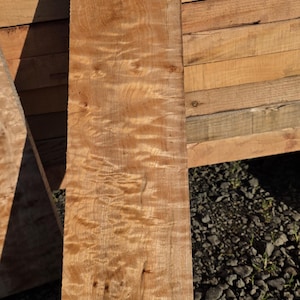 May include: A single, unfinished, rectangular piece of wood with a light brown color and a unique, wavy grain pattern. The wood appears to be a hardwood, possibly oak, and is ready for use in woodworking projects. The wood is approximately 60 inches long.