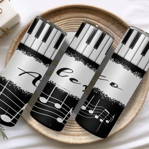 Könnte beinhalten: Drei schwarz-weiße Edelstahlbecher mit einem Klaviertasten-Design und Noten. Die Becher sind mit "Alto", "Tenor" und "Soprano" beschriftet.
