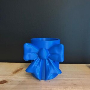 May include: A cylindrical blue cup featuring a prominent, three-dimensional bow attached to the front exterior. The entire object is a solid, uniform shade of blue with a smooth finish, showcasing a distinct bow shape with flowing ribbons.