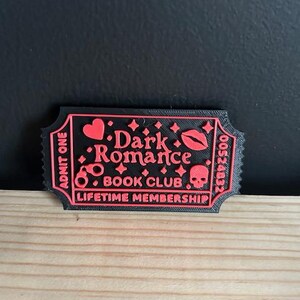 May include: A black and red ticket-shaped item with the words "Dark Romance Book Club" and "Lifetime Membership." The ticket features a heart, lips, a skull, and other symbols. The text "Admit One" is also visible.
