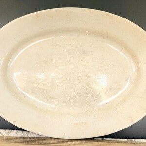 May include: Oval-shaped, off-white serving platter. The antique dish has a slightly raised rim and a smooth, glossy surface. The platter's color is a creamy, aged white, with subtle variations in tone across the surface. A classic piece for serving food.