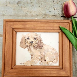 May include: A framed watercolor painting of a light brown cocker spaniel puppy sitting with its head tilted to the side. The painting is in a simple wooden frame.