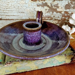 May include: A handcrafted ceramic candle holder with a deep purple and blue glaze. The holder features a central cup for the candle and a wide, shallow dish. The rim and handle are a darker shade of purple.