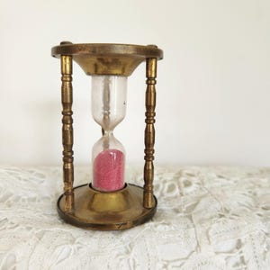 May include: An hourglass featuring a brass frame with four turned pillars connecting a circular top and base. The central glass bulb contains pink sand, with the majority of the sand settled in the bottom chamber of the clear glass vessel.