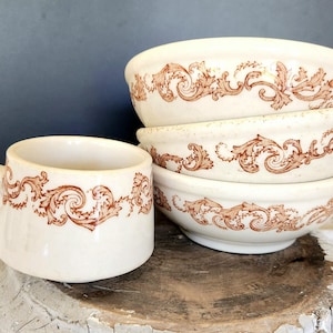 May include: A cream-colored ceramic mug with a handle and four matching bowls. Each piece features a brown, ornate scroll design around the rim. The mug and bowls are stacked on a wooden surface.