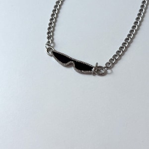 Może przedstawiać: Srebrny naszyjnik z wisiorkiem w kształcie okularów przeciwsłonecznych. Wisiorek w kształcie okularów przeciwsłonecznych jest czarny ze srebrnym obrysem i małymi, przezroczystymi zdobieniami. Łańcuszek ma wzór łańcucha, na białym tle.